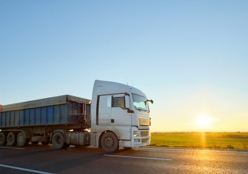 Semi-truck with tipping cargo trailer transporting sand from quarry driving on highway hauling goods in evening. Delivery transportation and logistics concept.