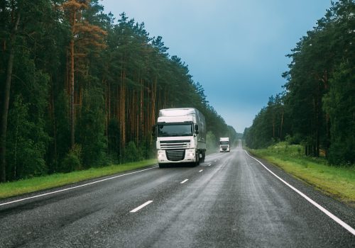 White Truck Or Tractor Unit, Prime Mover, Traction Unit In Motion On Road, Freeway. Asphalt Motorway Highway Against Background Of Forest Landscape. Business Transportation And Trucking Industry